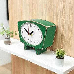 May include: A green wooden table clock with a white face, black hour and minute hands, and a red second hand. The clock features a trapezoidal shape with two short legs and black numerical markings for 12, 3, 6, and 9 on the dial.