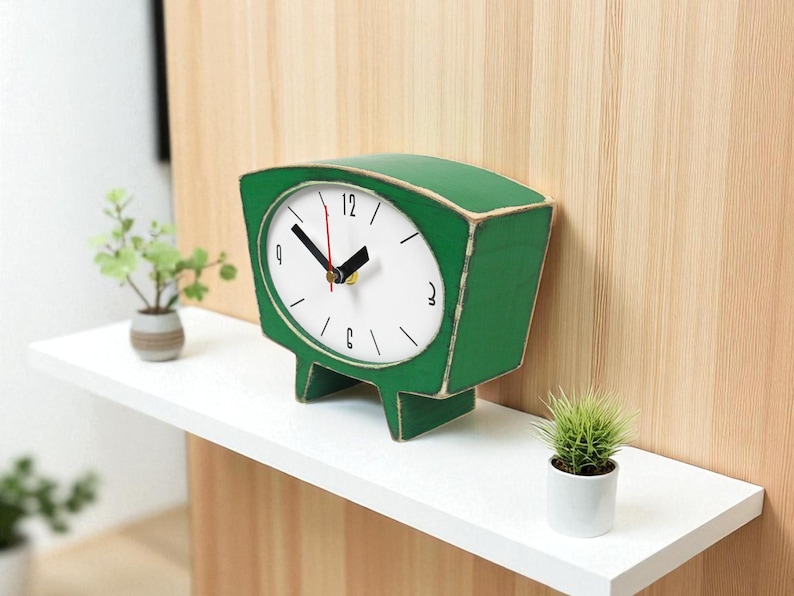 May include: A green wooden table clock with a white face, black hour and minute hands, and a red second hand. The clock features a trapezoidal shape with two short legs and black numerical markings for 12, 3, 6, and 9 on the dial.