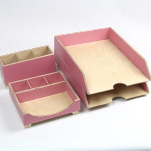 Op de afbeelding: Een set van drie houten bureau-organizers in lichtroze. De set bevat een rechthoekige pennenhouder met drie compartimenten, een vierkante organizer met vier compartimenten en een lade met twee niveaus. De interieurs zijn van natuurlijk hout.