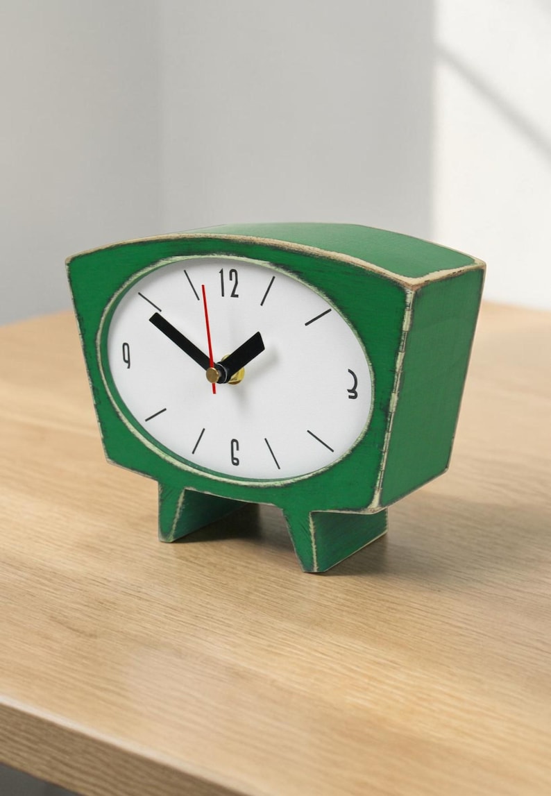 May include: A green table clock with a distressed finish and a white face. The clock features black hour and minute hands, a thin red second hand, and black numerals at the 12, 3, 6, and 9 positions. It stands on two short, wide legs.