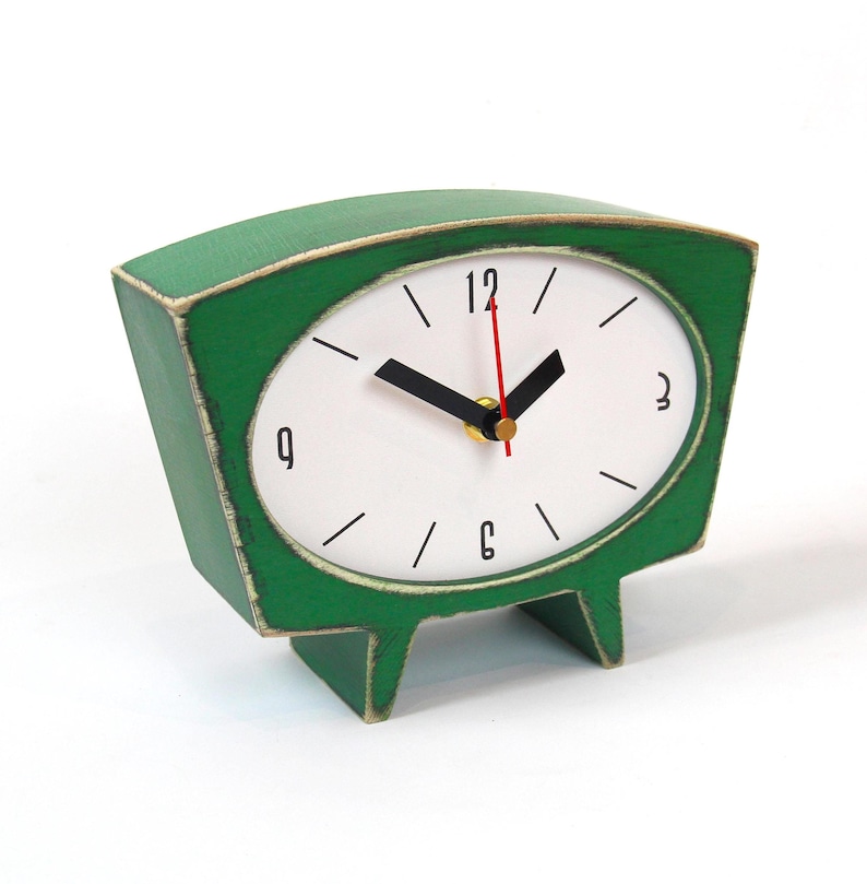 May include: A green table clock with a trapezoidal wooden frame featuring a distressed finish. The white oval clock face displays black numerals at 12, 3, 6, and 9, with black hour and minute hands and a thin red second hand on a central gold pin.