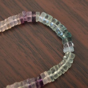Könnte beinhalten: Nahaufnahme eines Perlenarmbands mit Farbverlauf. Die Perlen sind quadratisch und gehen von Lila zu Grün über. Das Armband liegt auf einer dunkelbraunen Holzoberfläche.