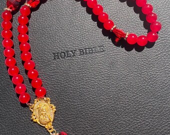 May include: A red beaded rosary with gold detailing and a gold pendant depicting a religious figure. The rosary is displayed on a black surface, with the words "HOLY BIBLE" visible.