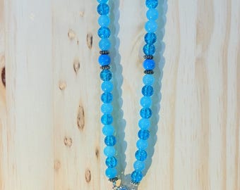 Puede incluir: Un rosario con cuentas azul claro y detalles plateados. El rosario presenta un colgante de cruz decorativo y una pieza central con una imagen religiosa. La cuenta superior es blanca con un diseño floral negro.