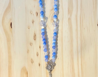 Puede incluir: Un rosario con cuentas azules y blancas, detalles de cristal transparente y un crucifijo plateado. El rosario presenta una cuenta decorativa de cerámica en la parte superior con un diseño floral.