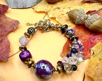 May include: A handcrafted bracelet featuring a mix of purple, gold, and clear beads. The bracelet includes a large, round, purple bead with a pearl-like finish, and smaller beads in various shapes and colors. The bracelet is set against a backdrop of autumn leaves.