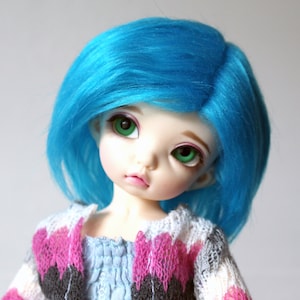 Könnte beinhalten: Eine Puppe mit leuchtend blauen Haaren und grünen Augen trägt einen rosa, weißen und grauen Pullover.