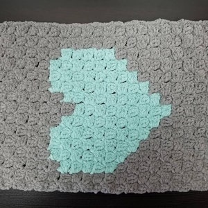 May include: A gray and teal crocheted rug with a unique design. The rug is approximately 12 inches by 18 inches.