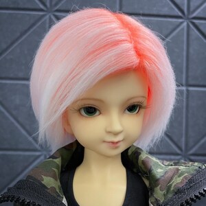 May include: A doll with short, pink and white hair, wearing a black tank top and a camouflage jacket.