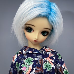 May include: A doll with white and blue hair wearing a blue hoodie with a colorful fish and lotus flower pattern.