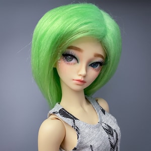 Puede incluir: Una muñeca con cabello verde y un top gris sin mangas con diseños de plumas negras. La muñeca tiene piel clara y ojos azules.