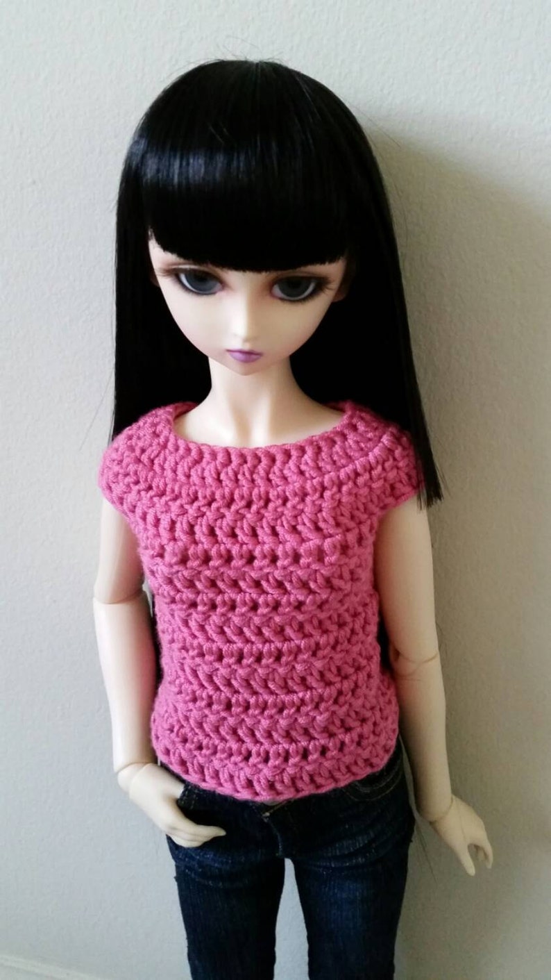 May include: A doll wearing a pink crocheted top with short sleeves and blue jeans.
