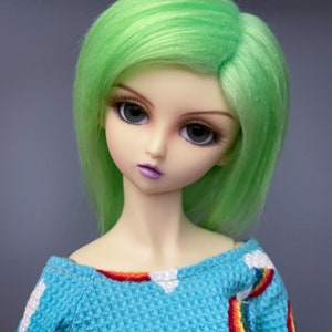 Puede incluir: Una muñeca con el pelo verde brillante que lleva una camisa azul y blanca con diseños de arcoíris.