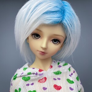 May include: A doll with white and blue hair wearing a white hooded sweater with a green, red, and purple pattern.