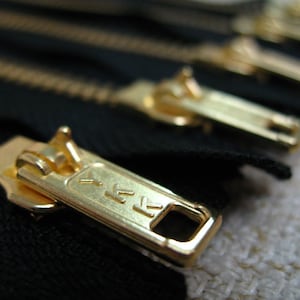 Könnte beinhalten: Nahaufnahme eines goldenen Reißverschluss-Schiebers mit dem Buchstaben "K" darauf. Der Reißverschluss-Schieber ist an einem schwarzen Stoffreißverschluss befestigt.