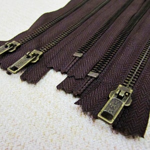 May include: Five brown zipper closures with brass teeth and sliders. The zippers are arranged in a row, with the sliders facing the viewer. The zippers are all the same size and style.