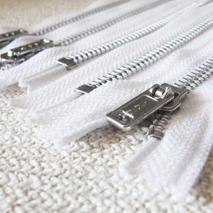Puede incluir: Primer plano de varias cintas de cremallera blancas con dientes metálicos plateados y un deslizador plateado. Las cremalleras están dispuestas sobre una superficie blanca texturizada.