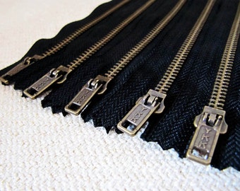 May include: Close-up of five black zipper tapes with gold metal teeth and YKK branded sliders. The zippers are arranged in a row, with the sliders facing the viewer.