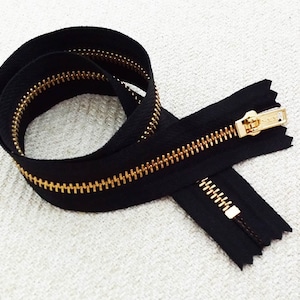 Puede incluir: Una cremallera negra con dientes dorados. La cremallera está enrollada y el tirador es visible.