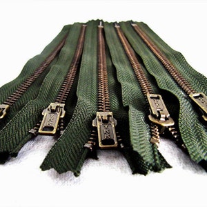May include: A close-up of six green zipper tapes with brass sliders. The zippers are arranged in a row, with the sliders facing the viewer. The zippers are all the same length and have a smooth, even texture.