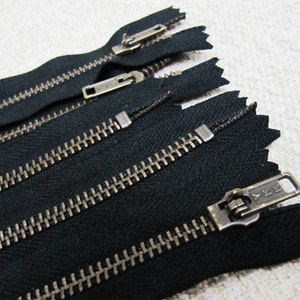 Pode incluir: Quatro fitas de zíper pretas com dentes e puxadores de metal prateado. Os puxadores do zíper têm o texto "YKK".