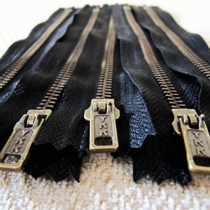 May include: Five black zipper tapes with brass YKK sliders. The zippers are laid out in a row, with the sliders facing the viewer.