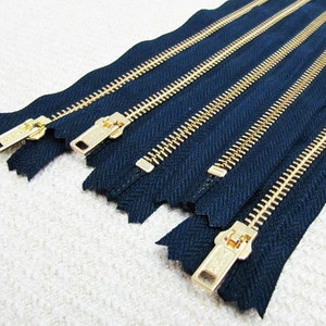 May include: Five gold metal zippers with navy blue tape. The zippers are arranged in a row, with the top zipper being the longest and the bottom zipper being the shortest.