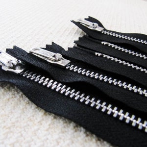 May include: Close-up of five black zipper tapes with silver teeth. The zippers are arranged in a row, with the top zipper slightly offset from the others.