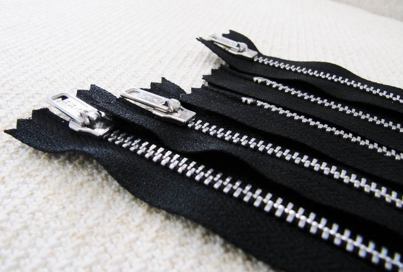 May include: Close-up of five black zipper tapes with silver teeth. The zippers are arranged in a row, with the top zipper slightly offset from the others.