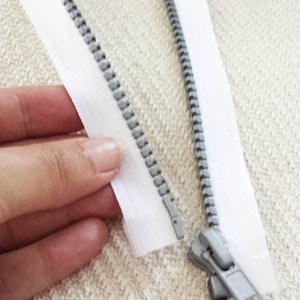 Puede incluir: Una cremallera blanca con un deslizador y dientes plateados. La cremallera está sobre una tela blanca texturizada.