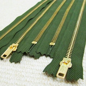 Puede incluir: Un conjunto de cinco cintas de cremallera verdes con dientes de metal dorado. Las cremalleras están dispuestas en fila, con los dientes hacia arriba.