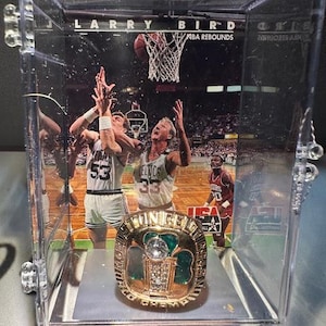 May include: A gold championship ring with a large, clear stone at its centre, displayed in a clear plastic case. The ring features green enamel accents and the words "UNICE" and "CHAMP" on its face. The case contains a background image of a basketball game with the name "LARRY BIRD" visible.
