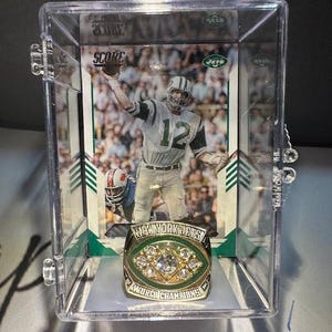 May include: A New York Jets World Champions ring with a clear plastic display case. The ring is gold with a green oval center and several clear stones. A football card with a player in a Jets uniform is behind the ring.
