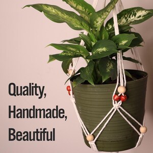 May include: A white macrame plant hanger with wooden beads and red accents holds a green potted plant. The text "Quality, Handmade, Beautiful" is visible in the image.