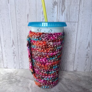 Puede incluir: Un colorido portavasos de ganchillo en tonos rosa, naranja, marrón y azul, envuelto alrededor de un vaso de plástico transparente con tapa azul y pajita amarilla. El portavasos tiene una correa para llevar.