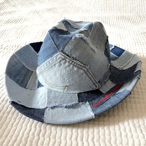 May include: A patchwork denim cowboy hat in shades of blue. The hat is constructed from various denim pieces, creating a unique, textured appearance. The brim and crown are stitched together, and the hat has a red embroidered detail.