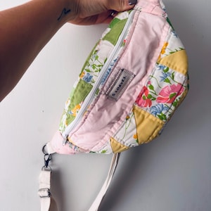 May include: A quilted fanny pack featuring a patchwork design of floral patterns in pink, green, and yellow. The bag includes a white zipper closure, an adjustable off-white strap with metal hardware, and a rectangular fabric label reading 5 LOAVES.