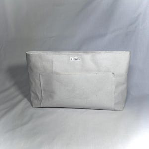 May include: An off-white tote bag with a rectangular shape and a large front pocket. The bag features the text "5 LOAVES" on the top. It is made from a textured fabric, and is designed for carrying items. The bag's simple design makes it versatile.