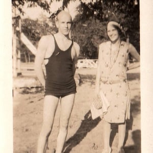 May include: Vintage black and white photograph of a man and a woman outdoors. The man wears a black swimsuit. The woman wears a patterned dress and a headband. They are standing on a grassy area.