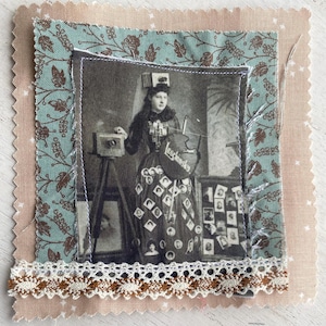 May include: A fabric square with a vintage black and white photograph of a woman with a camera. The image is framed with patterned fabric in shades of blue, brown, and pink, and trimmed with lace. The woman's dress is decorated with small photos, and the text "Leschnsky" is visible.