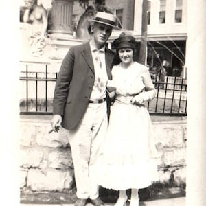 May include: Black and white photograph of a couple posing outdoors. The man wears a suit jacket, light-colored pants, and a hat. The woman is in a dress and hat. A stone fountain and building are in the background.