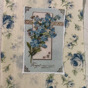 May include: A rectangular fabric patch featuring a cream background with a blue floral pattern. Centered on the patch is a smaller, rectangular fabric piece with a light blue border, displaying blue forget-me-not flowers and the text Forget me not.