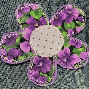May include: A fabric flower applique featuring five petals with a purple and green floral print on a black background. The center is a light lavender circle with small brown dots. The edges are finished with a white zigzag stitch.