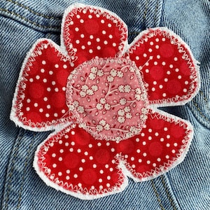 May include: A handmade fabric flower applique with a red polka dot pattern and a floral center. The flower is stitched onto a denim background, showcasing a charming, handcrafted design. The applique has a white stitched border.