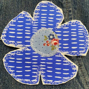 May include: A fabric flower applique featuring five petals made of blue fabric with a white dash pattern. The center is a circular light blue patch with a small floral print of red and blue flowers. The edges are finished with white zigzag stitching.