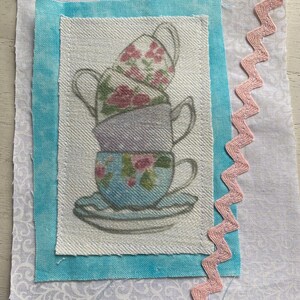 Könnte beinhalten: Ein Stoffquadrat mit einem Design aus gestapelten Teetassen mit floralen Mustern in Rosa, Blau und Lila. Die Tassen befinden sich auf einem weißen Hintergrund, umrahmt von einem hellblauen Rand und einer rosa Zickzack-Borte.