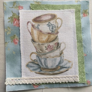 Könnte beinhalten: Ein Stoffquadrat mit einem Aufdruck von gestapelten Vintage-Teetassen in Beige, Blau und Rosa. Die Tassen befinden sich auf einem weißen Hintergrund, der mit floralem und grünem Stoff überlagert und mit Spitze verziert ist. Das Design hat eine Vintage-Ästhetik.