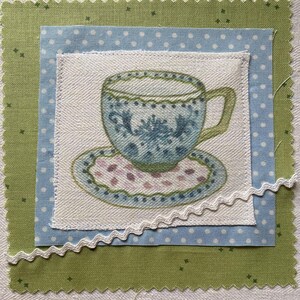 Könnte beinhalten: Ein Stoffquadrat mit einer detaillierten Illustration einer Teetasse und Untertasse in Blau-, Grün- und Rosatönen. Das Teetassen-Design befindet sich auf einem weißen Quadrat, das über einem blauen Quadrat mit Punkten und einem grünen Quadrat mit gewelltem Rand liegt.