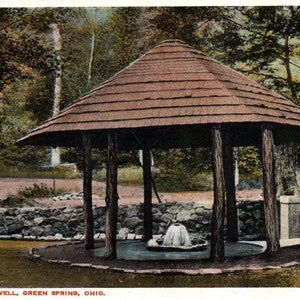 Könnte beinhalten: Ein hölzerner Pavillon mit einem Schindeldach schützt eine natürliche Quelle mit einem kleinen Springbrunnen. Eine rote Holzbank befindet sich rechts vom Pavillon. Der Text "ARTESIAN MINERAL WELL, GREEN SPRING, OHIO." ist unter dem Bild gedruckt.