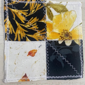 May include: Square fabric coaster with a patchwork design. The coaster features four squares with different patterns: black and gold leaves, yellow flowers, white with orange leaves, and black with dark leaves. White zigzag stitching outlines the edges.